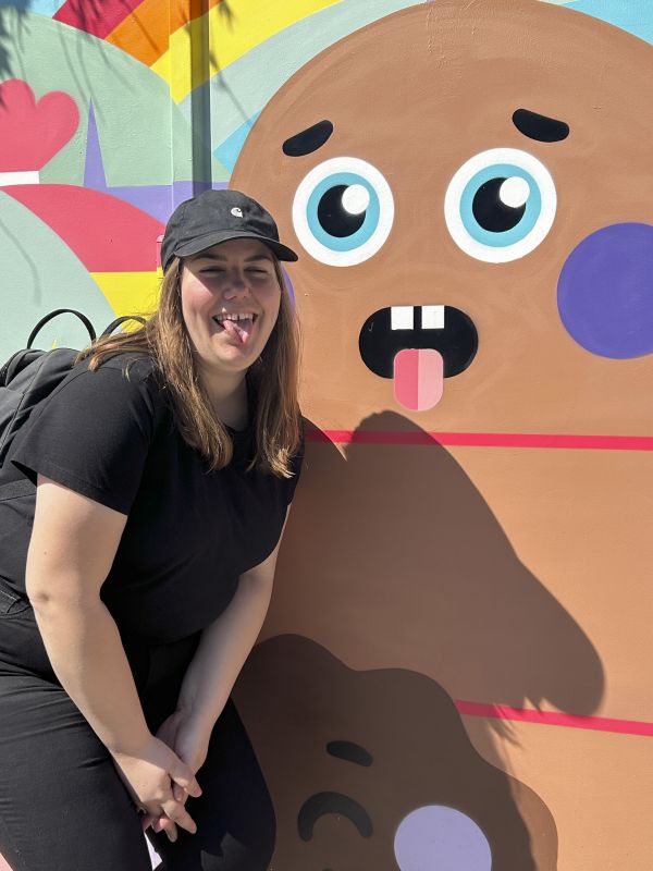 Roberta posiert vor einer Wandmalerei mit einer Figur, die die Zunge rausstreckt